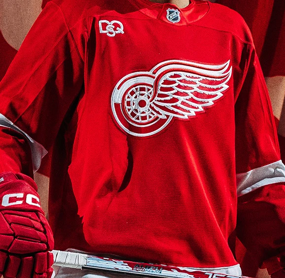Man wearing red Detroit Red Wings jerseys and ball cap standing inside of the hockey arena.
