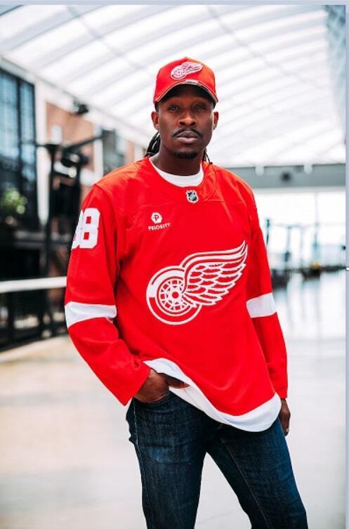 Man wearing red Detroit Red Wings jerseys and ball cap standing inside of the hockey arena.