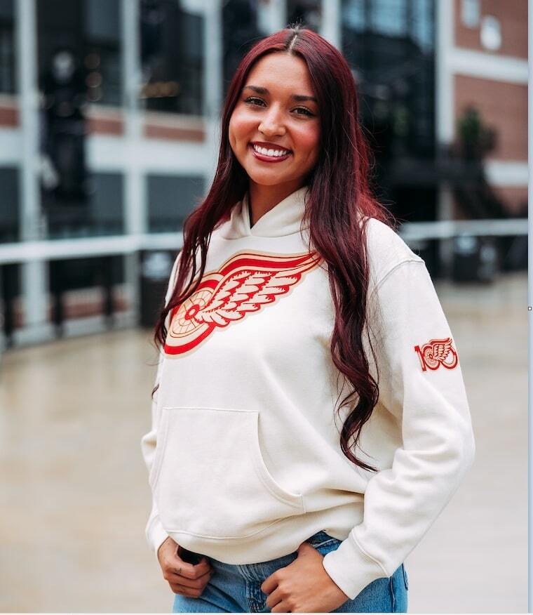 Woman smiling and standing inside arena wearing a white Detroit Red Wings pullover hooded sweatshirt with a vintage winged wheel logo on front and centennial logo on left sleeve.