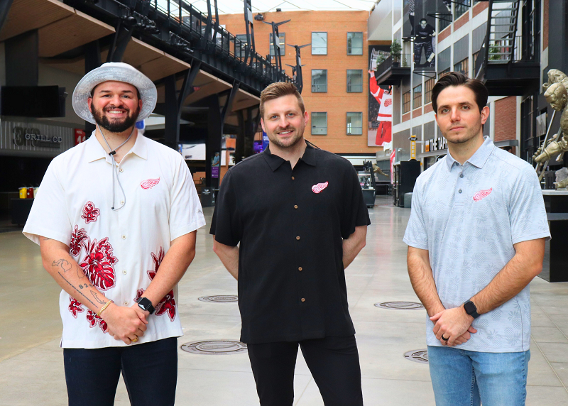 Three men posing outside of the Detroit Red Wings hockey arena wearing Detroit Red Wings button up shirts in three different styles.
