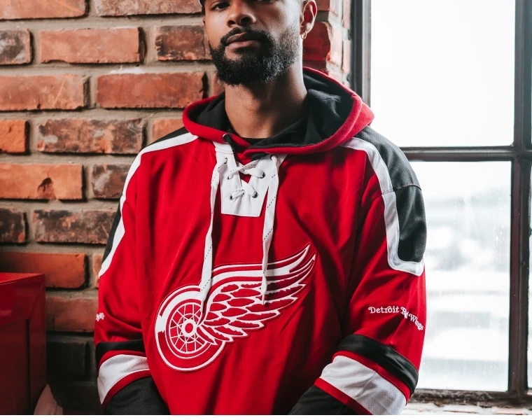 Man leaning against rail inside arena wearing a red pullover hooded sweatshirt with Detroit Red Wings vintage winged wheel logo on front. 