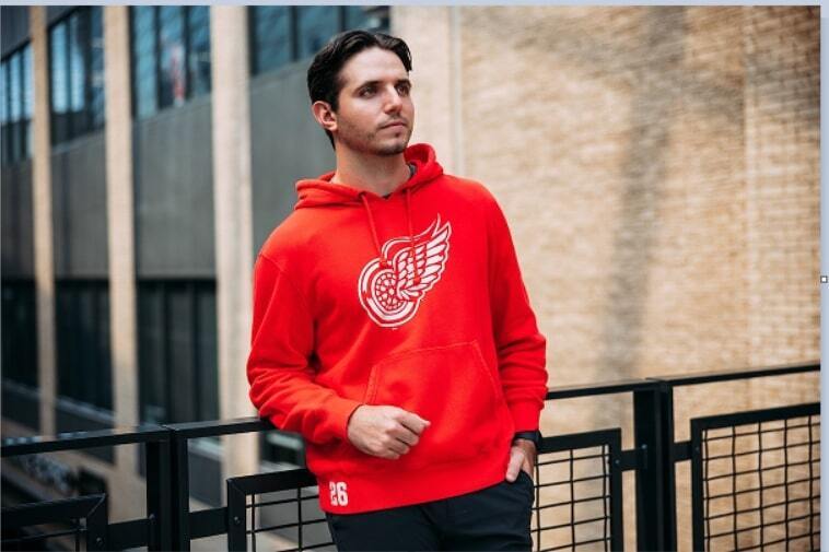 Man leaning against rail inside arena wearing a red pullover hooded sweatshirt with Detroit Red Wings vintage winged wheel logo on front. 