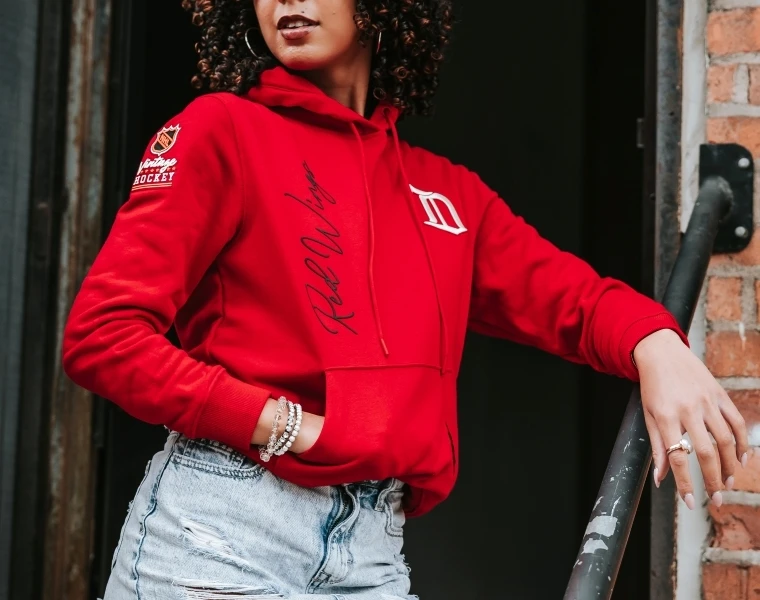 Woman smiling and standing inside arena wearing a white Detroit Red Wings pullover hooded sweatshirt with a vintage winged wheel logo on front and centennial logo on left sleeve.